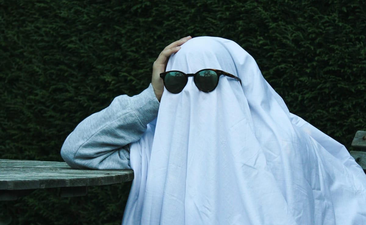 Person in sweater with sheet over their head and most of body wearing black oval sunglasses looking like a cool casual ghost