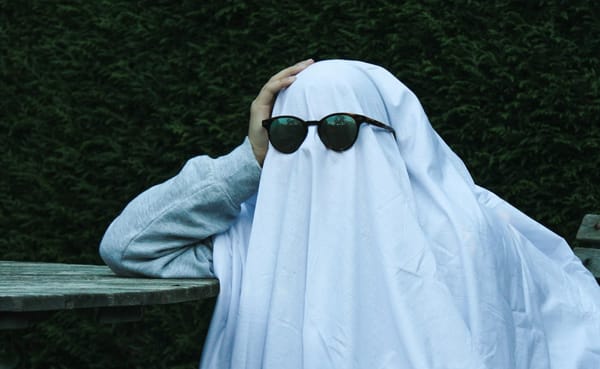 Person in sweater with sheet over their head and most of body wearing black oval sunglasses looking like a cool casual ghost